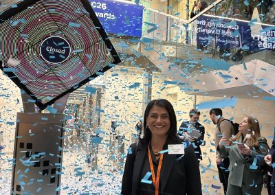 Tab Ahmad OBE, wearing a black blazer and an event lanyard, smiles warmly as blue and white confetti falls all around her. To her left is a large, angled digital display showing the word "Closed" within colorful, concentric rings, resembling a stock exchange closing event. In the background, people can be seen watching and taking photos from the glass balconies of a modern, multi-level atrium.